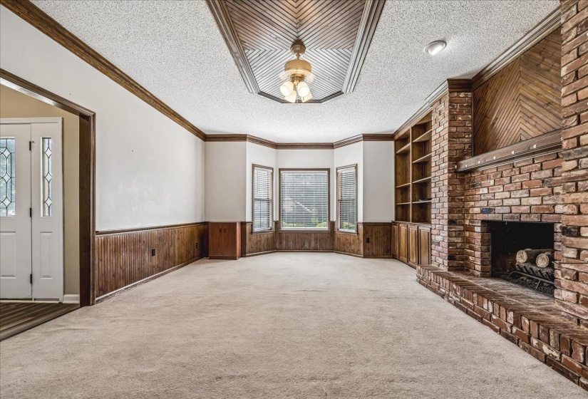 Spacious living area featuring a prominent brick fireplace with hearth, built-in shelving, bay windows, wood paneling, and a decorative ceiling with a central lighting fixture