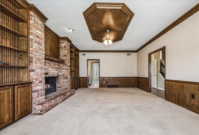 Spacious living area featuring a brick fireplace with a raised hearth, built-in wood cabinetry and shelving, wood paneled walls, and a wood-paneled octagon tray ceiling