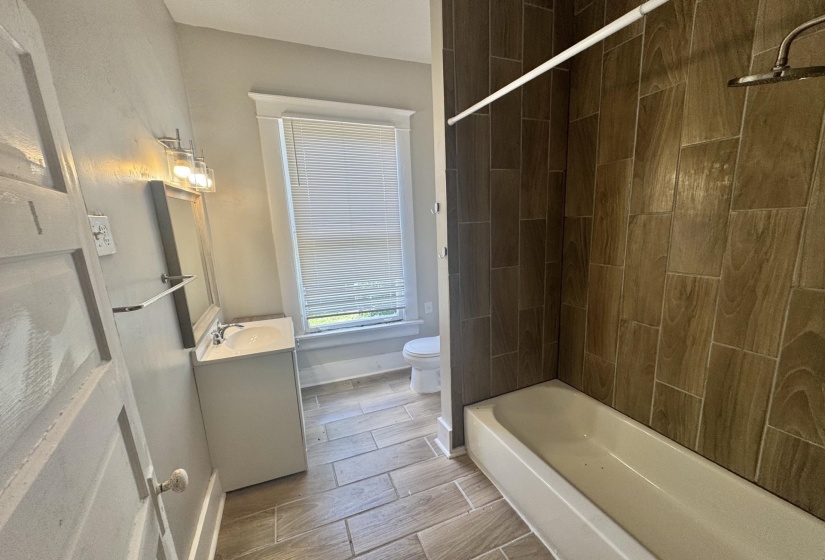Bathroom featuring a full tub with a tile surround, wood-finish tile flooring, a single vanity with a basin sink, and a window with blinds