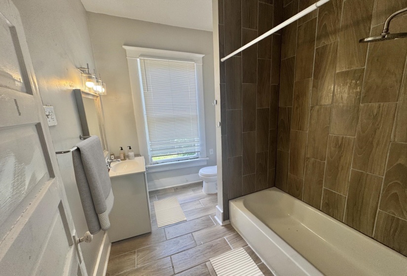 Bathroom featuring a tiled shower surround, a white bathtub, a single vanity with storage, a wall-mounted mirror, and wood-finish tile flooring