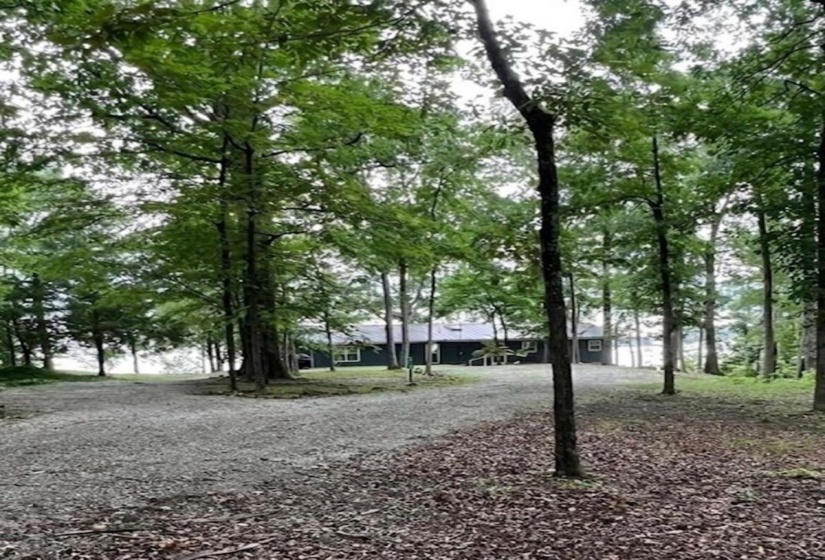 Gravel driveway bordered by mature trees, leading to a dark-toned residence with a visible body of water in the background