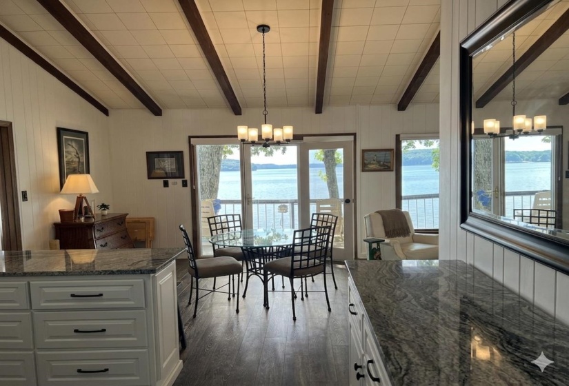 Open-concept interior featuring a breakfast bar with a stone-finish countertop, wood-finish flooring, exposed ceiling beams, and a dining area with direct access to an outdoor deck