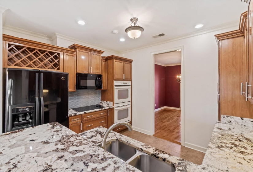 Kitchen with brown cabinetry, black appliances, light stone countertops, ornamental molding, and light tile patterned floors