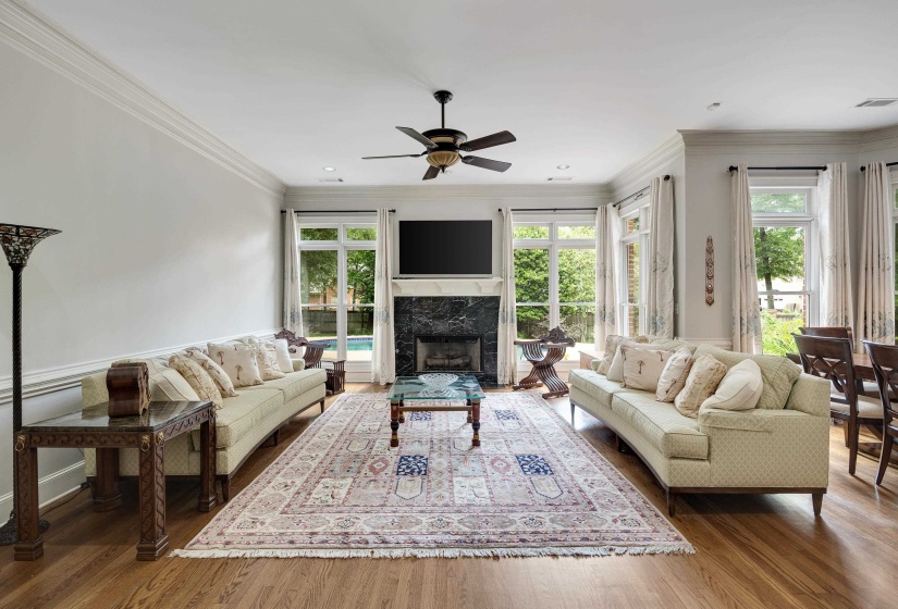 Bright living room with hardwood flooring, a black marble fireplace, extensive crown molding, a ceiling fan, and large windows