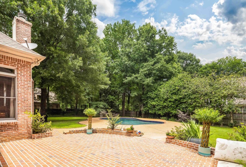 Expansive brick patio featuring a curved retaining wall, a kidney-shaped swimming pool, mature perimeter trees, and a well-maintained lawn