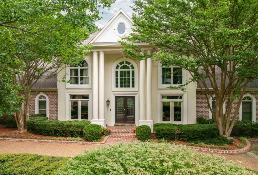 Stately residence featuring a prominent portico with four columns, a double-door entry, a symmetrical facade with arched and rectangular windows, a brick accent wall, and a gabled roofline with an oval window