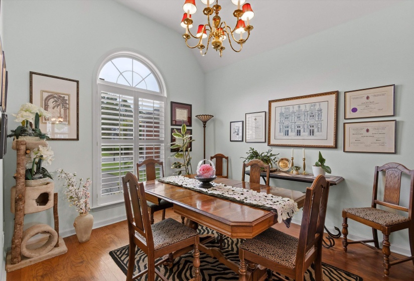 Vaulted ceiling dining area featuring a prominent arched window with white plantation shutters, hardwood flooring, and a brass chandelier