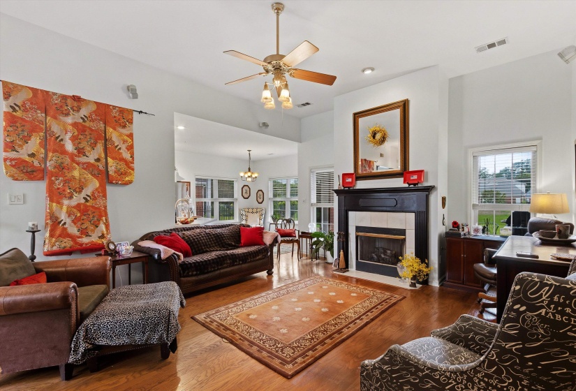 Living area featuring high ceilings, wood-finish flooring, a ceiling fan with integrated lighting, a fireplace with a dark wood mantel and white tile surround, and multiple windows