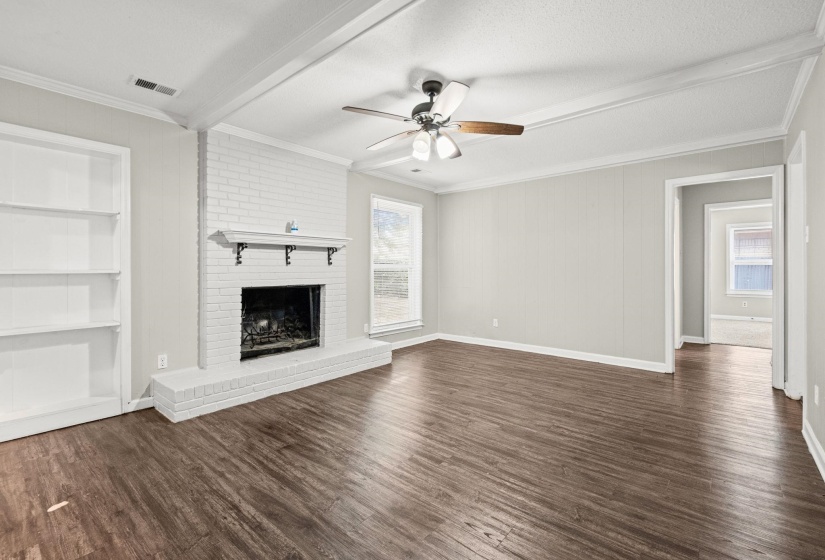 Spacious living area featuring wood-finish flooring, a white brick fireplace with mantel, built-in shelving, a ceiling fan, and crown molding