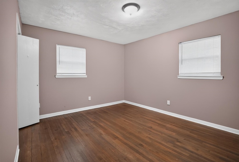 Spacious interior room featuring wood-finish flooring, two windows with blinds, a white door, white baseboards, and a ceiling-mounted light fixture