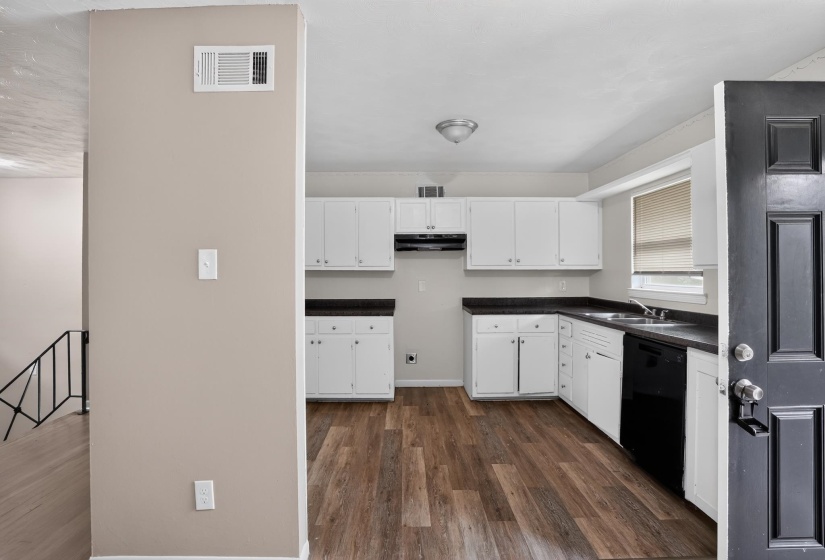 The kitchen features wood-finish flooring, white cabinetry, dark countertops, and a black dishwasher