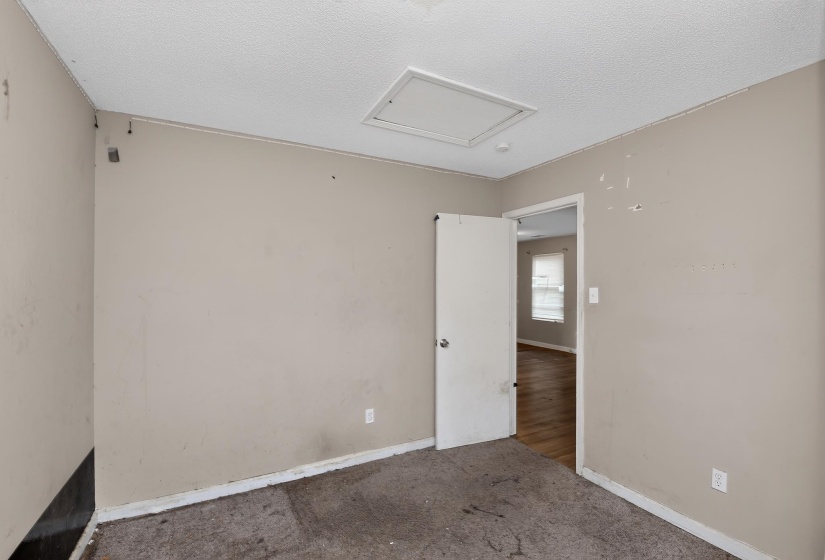Neutral-toned walls frame an interior room with a ceiling-mounted access panel and a white door leading to an adjacent area featuring wood-finish flooring and a window