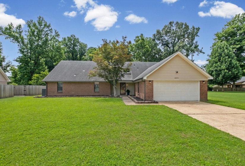 Brick-front residence featuring an attached garage, paved driveway, mature trees, and a spacious front lawn