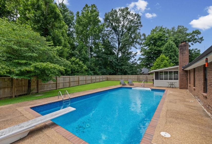 Expansive backyard featuring a rectangular swimming pool with diving board and integrated steps, a surrounding concrete patio, a privacy fence, and a brick exterior home with a sunroom addition