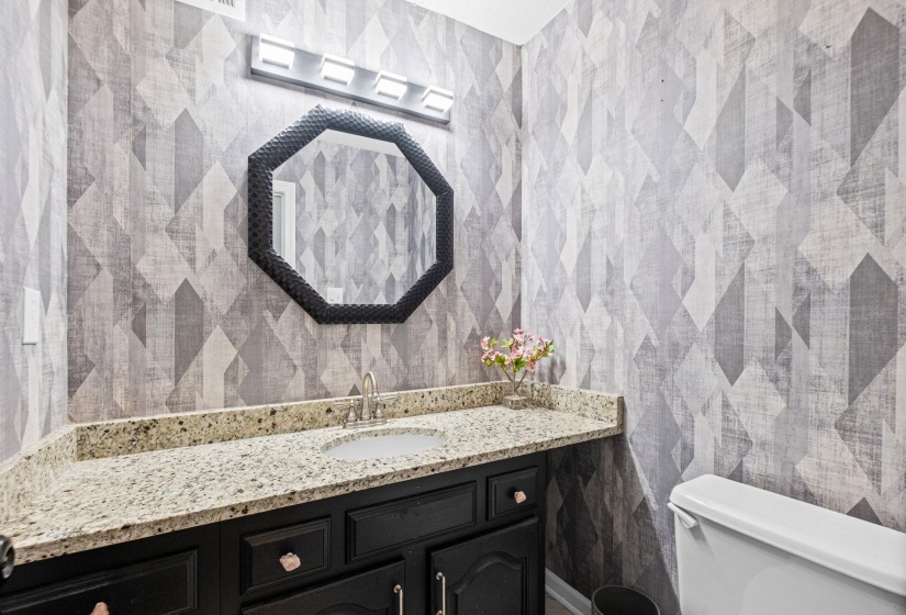 Bathroom featuring geometric pattern wallpaper, a dark wood-finish vanity with an integrated sink, a light-toned speckled countertop, and a contemporary octagonal mirror