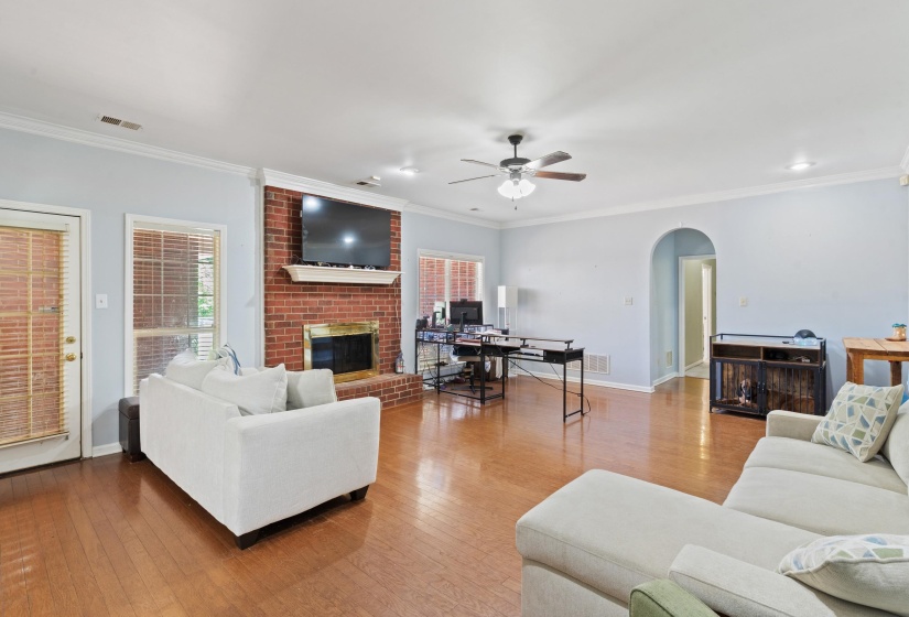 Spacious living area featuring wood-finish flooring, a brick fireplace with mantel, ceiling fan, and multiple windows