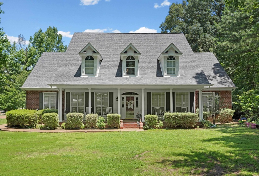 Brick facade residence featuring a covered front porch with white columns, symmetrical dormer windows, and a manicured lawn with established landscaping