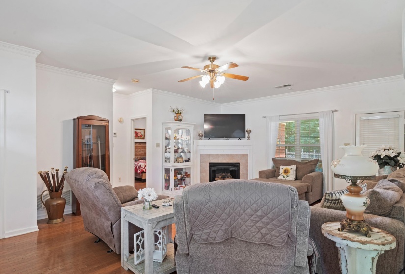 Living area featuring wood-finish flooring, a ceiling fan with integrated lighting, crown molding, and a fireplace with a light-colored mantel and surround