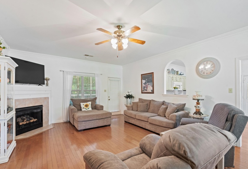 Living area featuring wood-finish flooring, a corner fireplace with stone surround, a ceiling fan, and crown molding