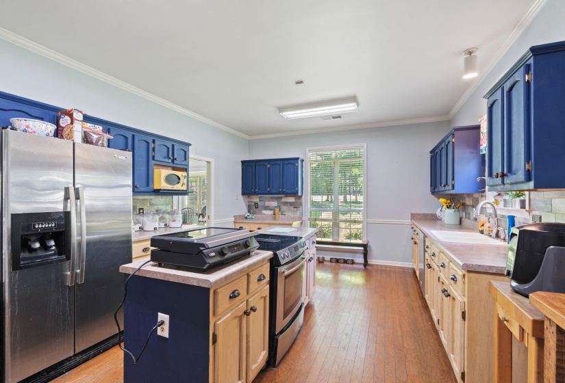 Spacious kitchen featuring wood-finish flooring, two-tone cabinetry with blue upper cabinets and light wood lower cabinets, light-colored countertops, stainless steel appliances, and a window overlooking exterior landscaping