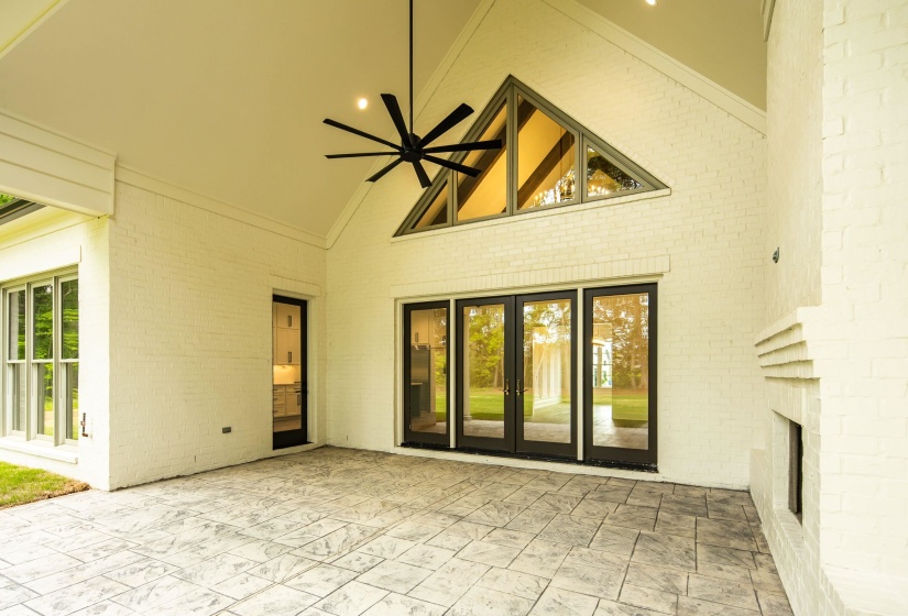 Covered patio featuring an expansive stamped concrete surface, a vaulted ceiling with an oversized ceiling fan, a prominent brick fireplace, and black-framed French doors