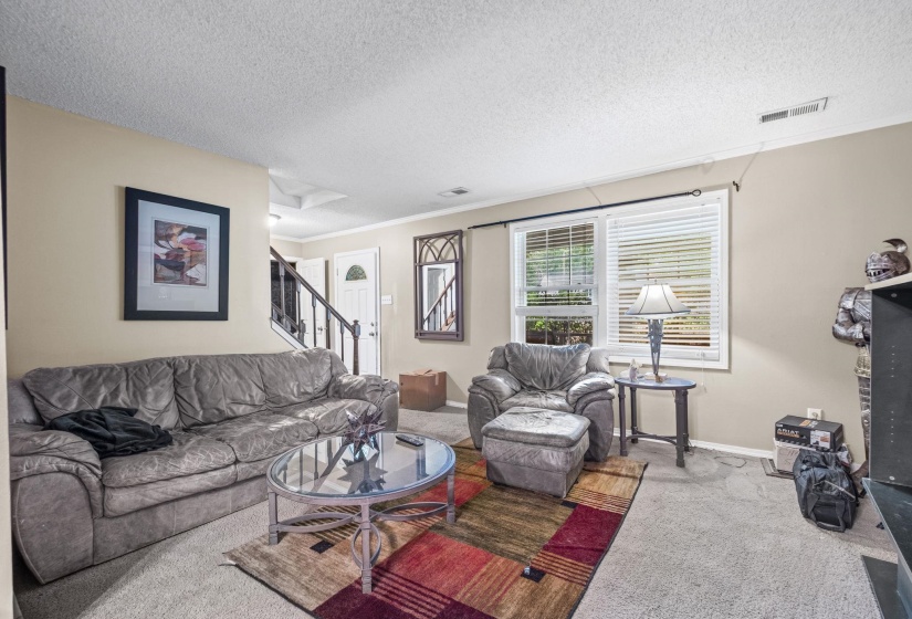 Carpeted living area featuring a large window with blinds, neutral wall tones, white trim, and a staircase with dark railings