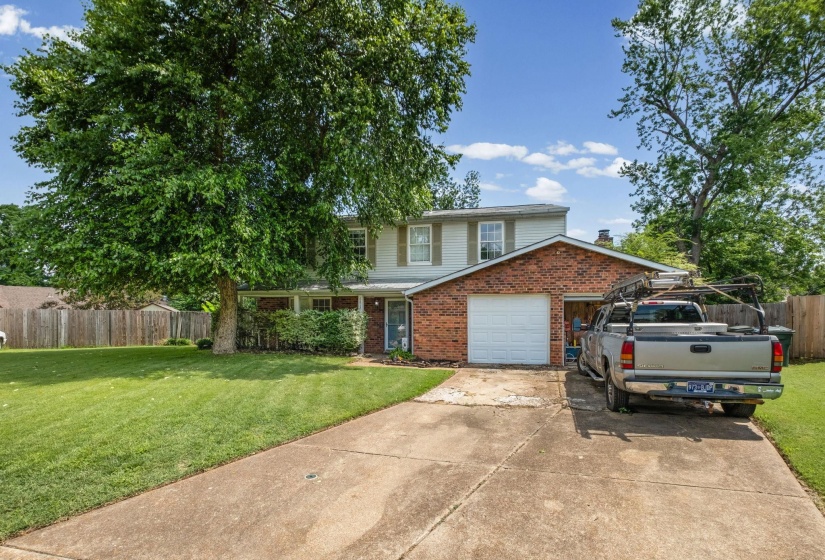 Two-story brick and siding residence featuring a two-car garage, mature front yard tree, concrete driveway, and established foundation plantings