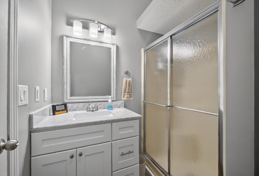 Bathroom featuring a single vanity with a white countertop and undermount sink, white shaker-style cabinetry, a framed mirror, and a shower with sliding textured glass doors
