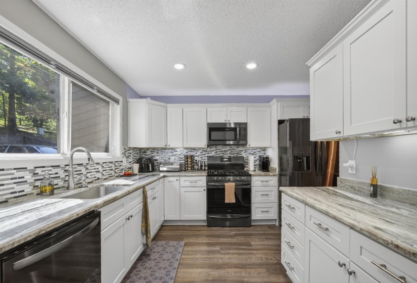 Kitchen featuring white shaker-style cabinetry, stainless steel appliances, mosaic tile backsplash, wood-finish flooring, and light-toned granite countertops