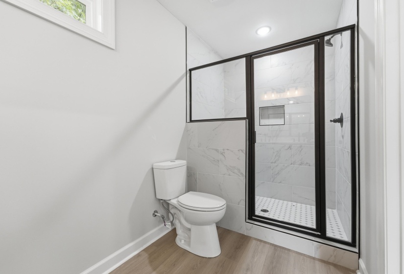 Contemporary bathroom featuring a frameless glass shower enclosure with matte black trim, white subway tile with a marble-effect, a built-in shower niche, a white toilet, and wood-finish flooring