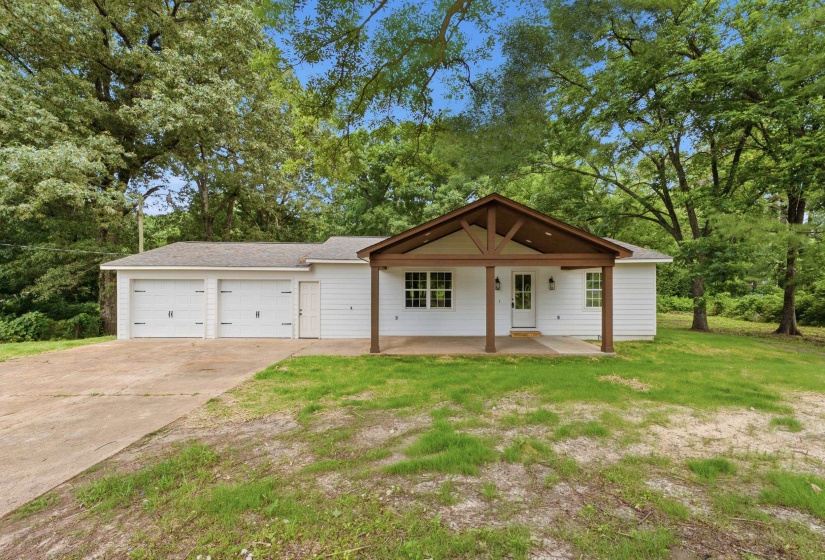 White-sided home featuring a covered front porch with exposed wood beams and columns, a two-car garage with carriage-style doors, a concrete driveway, and surrounding mature trees