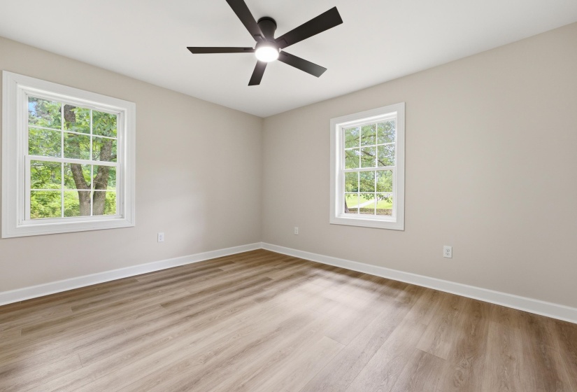 Spacious room featuring wood-finish flooring, two large pane windows with white trim, a ceiling fan with integrated lighting, and neutral wall tones