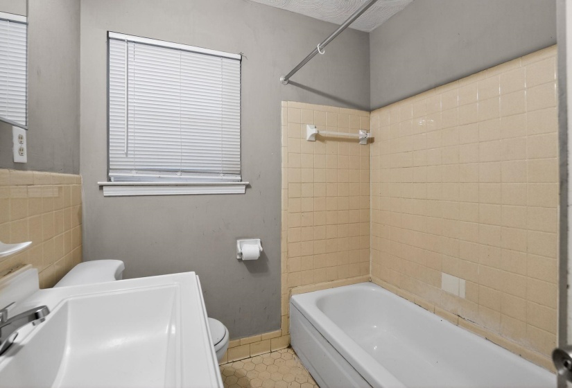 Bathroom featuring a window with blinds, a white vanity with an integrated sink, a commode, a bathtub with tiled surround, and hexagonal floor tiles