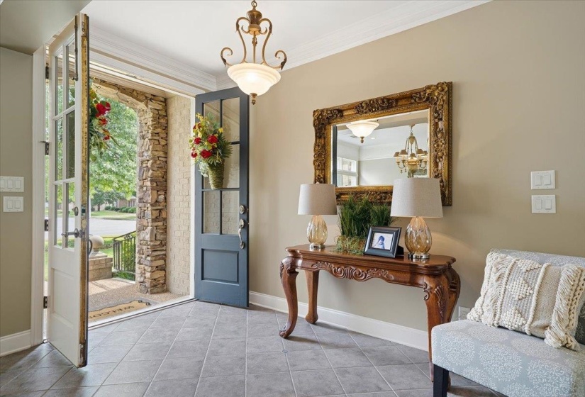 Welcoming entryway featuring dual glass-paneled doors, stone archway, tile flooring, an overhead brass chandelier, and crown molding