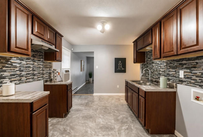 Kitchen featuring ample dark wood cabinetry, a mosaic tile backsplash, light-toned countertops, a double basin stainless steel sink with a gooseneck faucet, and durable tile flooring