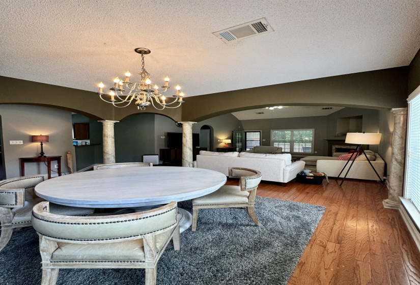 Spacious open-concept living area featuring a traditional multi-arm chandelier, decorative columns, arched doorways, and warm wood-finish flooring
