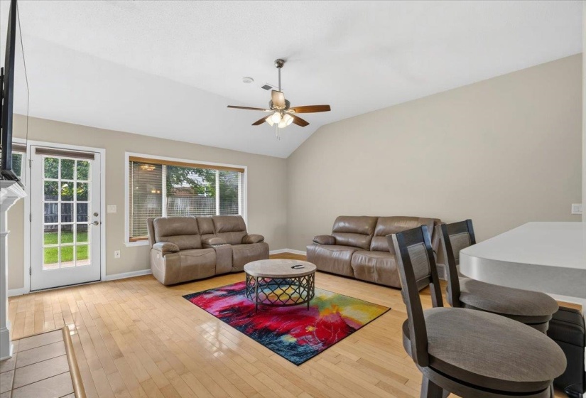 Living area featuring vaulted ceilings, wood-finish flooring, a ceiling fan, and abundant natural light from the large window and glass-paned door
