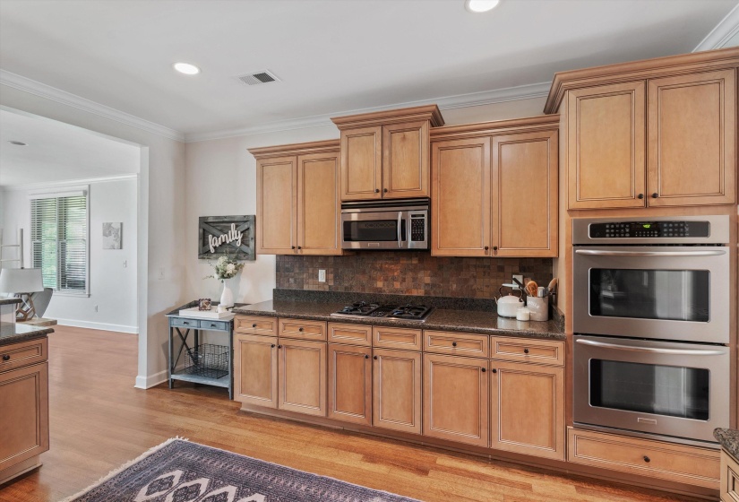 Kitchen featuring wood-finish flooring, light wood cabinetry with dark hardware, a dark stone countertop, a tile backsplash, and stainless steel appliances including a built-in microwave, gas cooktop, and double ovens
