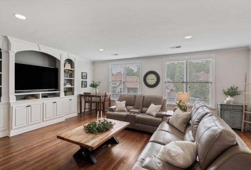 Expansive living area featuring rich wood-finish flooring, a built-in media center with integrated shelving and cabinetry, recessed lighting, and multiple windows with blinds