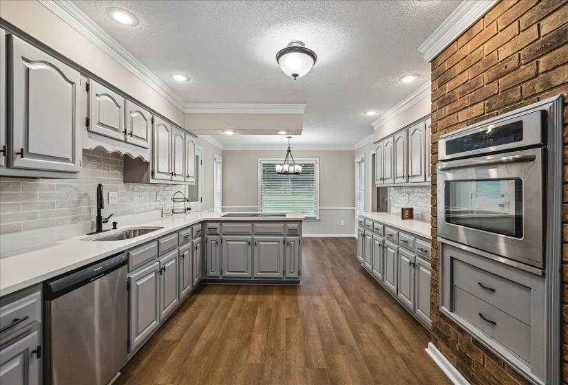 Expansive kitchen featuring light gray cabinetry, white countertops, wood-finish flooring, stainless steel appliances, and a brick accent wall with a built-in oven