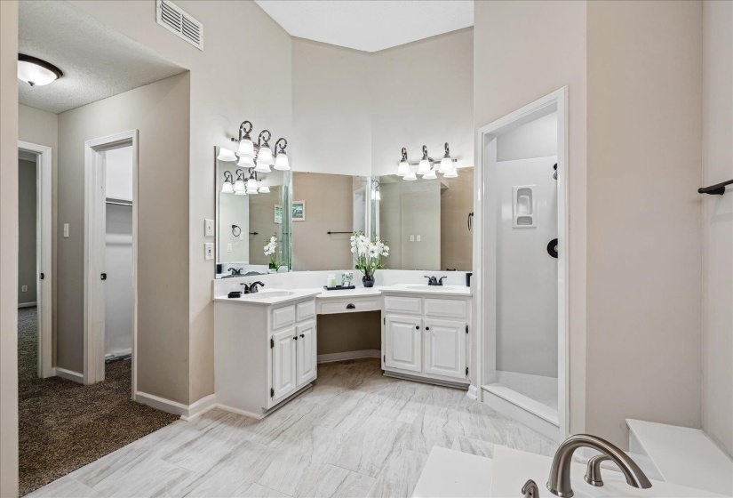 Expansive bathroom featuring dual vanities with white cabinetry, a built-in makeup vanity, a walk-in shower with a glass enclosure, and a built-in tub with a brushed nickel faucet