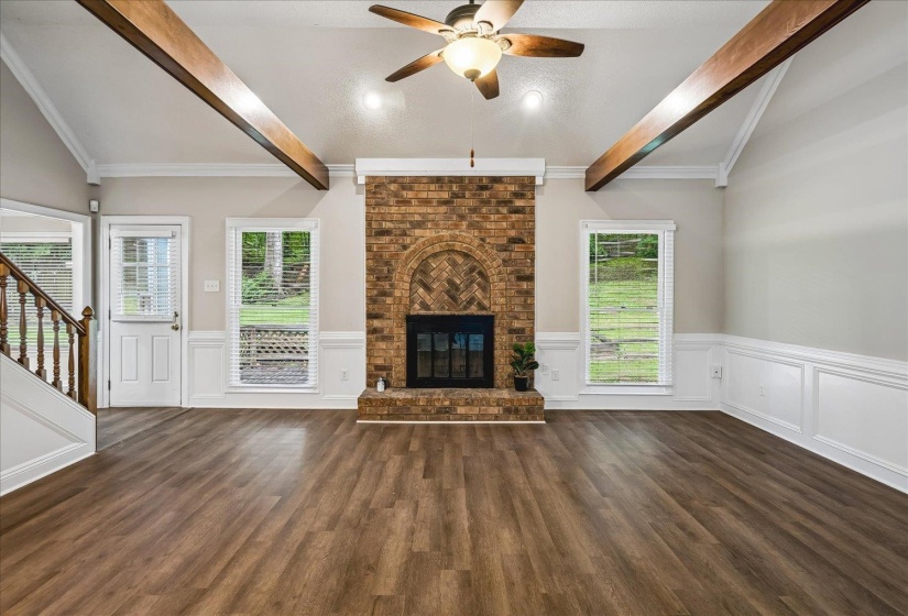Spacious living area featuring a brick fireplace with an arched hearth, vaulted ceilings with exposed wood beams, wood-finish flooring, and white wainscoting