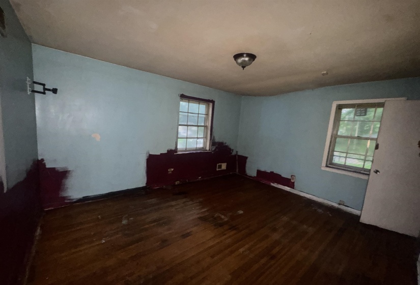 Room featuring wood-finish flooring, two window openings with visible exterior greenery, a ceiling-mounted light fixture, and a wall-mounted bracket