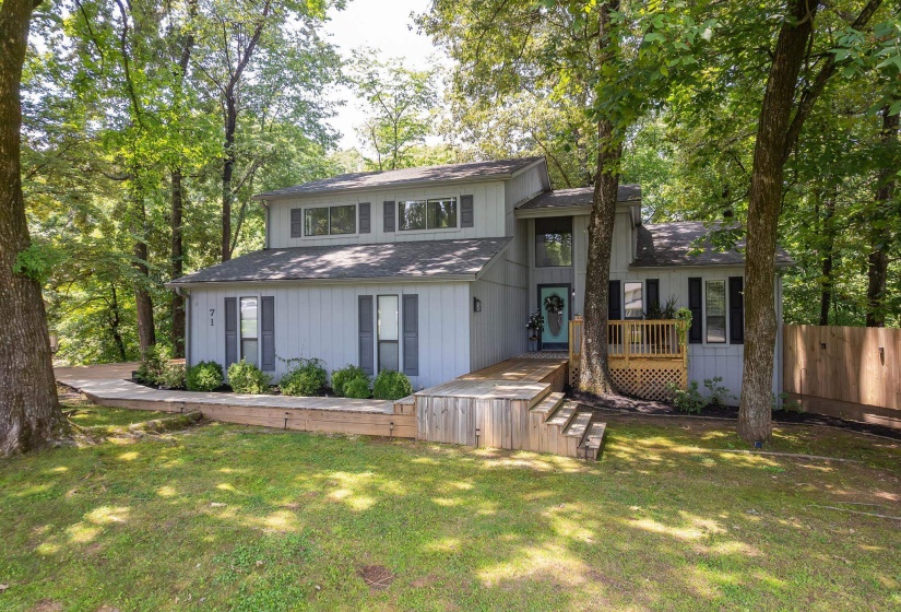 Contemporary exterior featuring a light gray facade, dark shutters, an inviting blue front door, a multi-level wooden deck, and mature tree landscaping