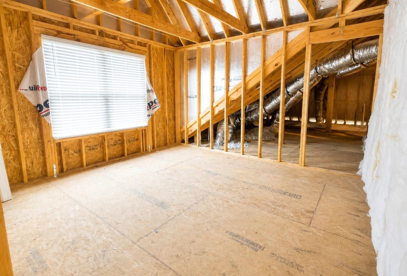 Unfinished room featuring exposed wood framing, plywood subflooring, and a single window with blinds