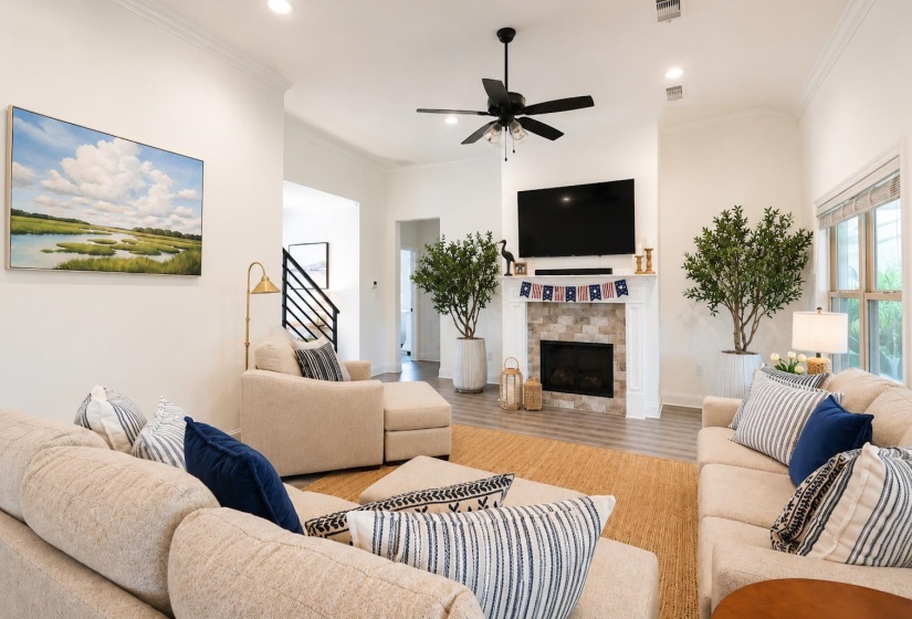 Spacious living area featuring a central fireplace with stone surround and white mantel, a ceiling fan, recessed lighting, wood-finish flooring, and crown molding
