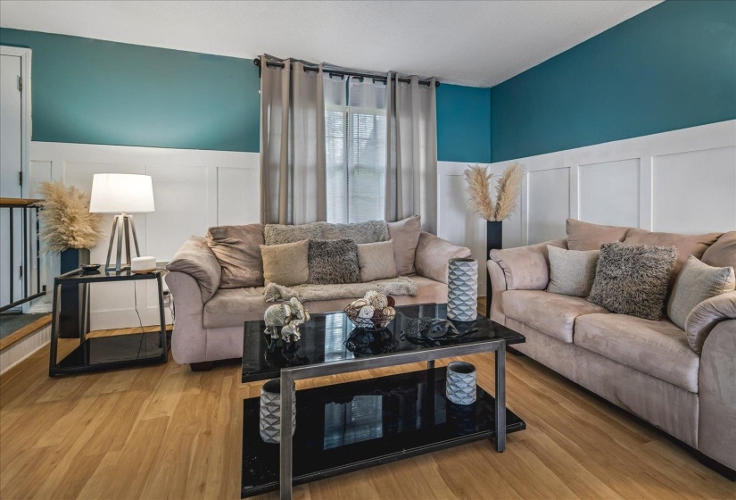 Living area featuring wood-finish flooring, wainscoting with a contrasting teal wall, and natural light from a large window with drapes