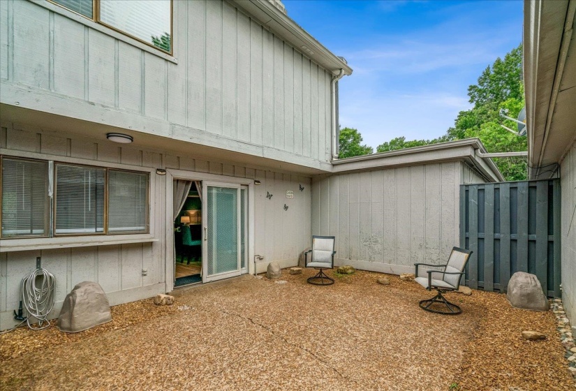 Private patio with a concrete surface and surrounding gravel, featuring a sliding glass door entry, multiple windows with blinds, and exterior lighting