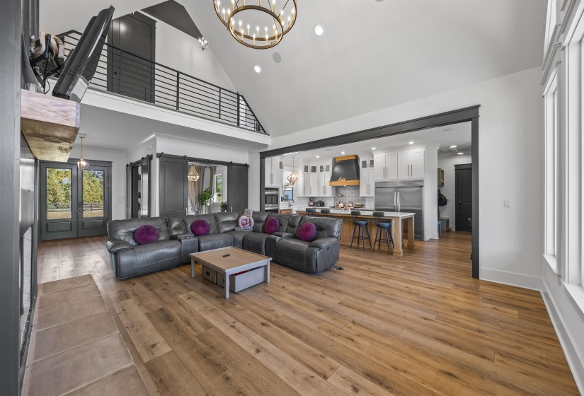 Expansive living area featuring vaulted ceilings, wood-finish flooring, a kitchen island with seating, a loft with metal railing, and glass-paned double doors