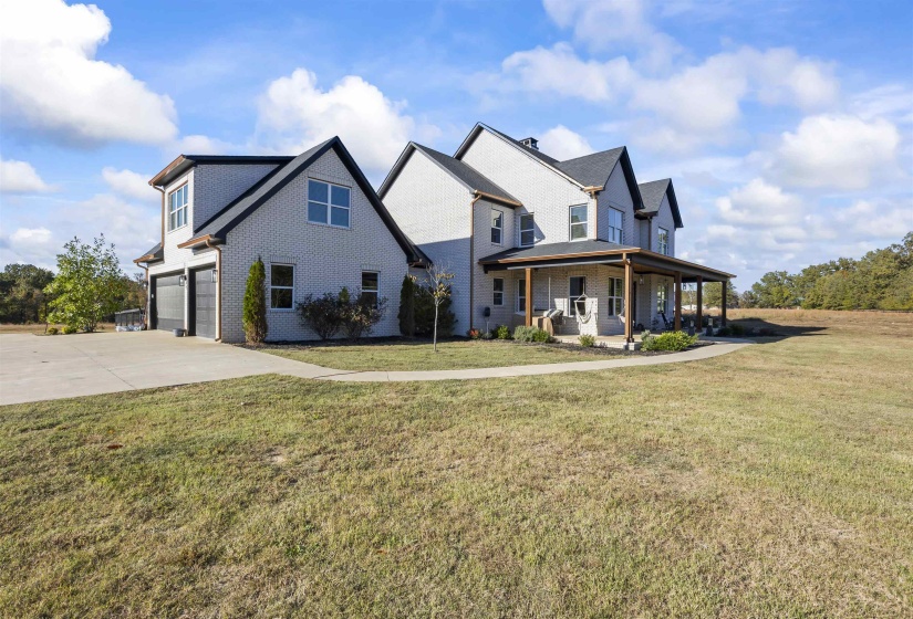 Two-story residence featuring light gray brick veneer, dark gabled roofing, a covered front porch with wood columns, an attached garage with dark doors, and a concrete driveway